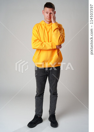Portrait of young man in yellow hoodie with face palm gesture on gray background 114503597