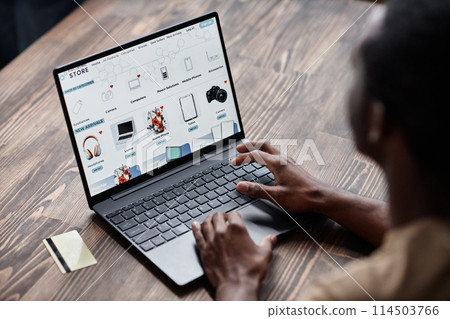 High angle view of unrecognizable Black man sitting at wooden desk choosing goods in online shop on laptop 114503766