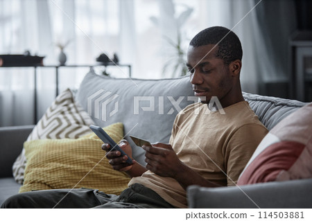 Young African American man sitting on sofa in living room paying for goods online using app on smartphone, copy space Young African American man sitting on sofa in living room paying for goods online using app on smartphone, copy space 114503881