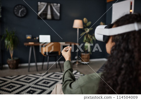 Over-the-shoulder shot of unrecognizable young woman wearing VR headset sitting in living room surfing Internet 114504009