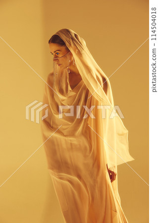 Muse. Beautiful young woman in flowing light fabric with face covered with golden accessories standing against sand color background 114504018