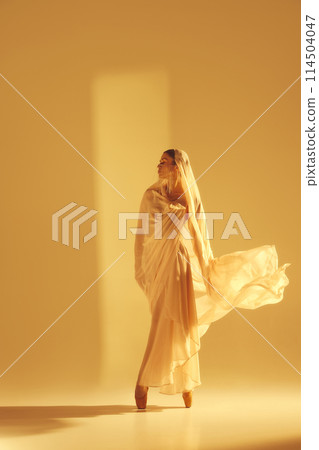 Ethereal beauty. Mystical scene of female ballet dancer with golden flowing fabric performing, symbolizing purity and elegance of ballet 114504047