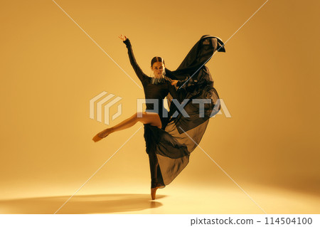 Artistic and expressive performance. Young woman, ballet dancer in black flowing attire dancing against golden background 114504100
