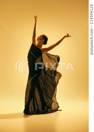Simplicity and beauty of the dance. Full-length image of elegant woman, ballerina in black costume dancing against yellow background 114504128