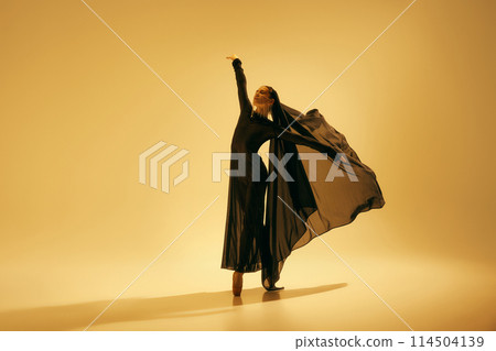 Desert freedom. Elegant and artistic young woman in black attire with veil dancing, performing ballet against sand color background Desert freedom. Elegant and artistic young woman in black attire with veil dancing, performing ballet against sand color background 114504139