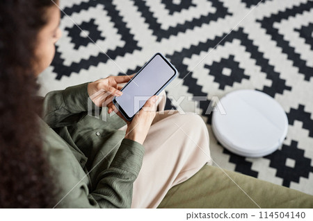 High angle view of unrecognizable young woman using robotic vacuum cleaner app on smartphone High angle view of unrecognizable young woman using robotic vacuum cleaner app on smartphone 114504140