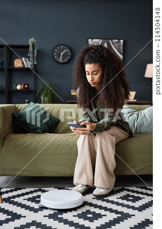 Vertical shot of young woman sitting in living room choosing robotic vacuum settings on smartphone 114504148
