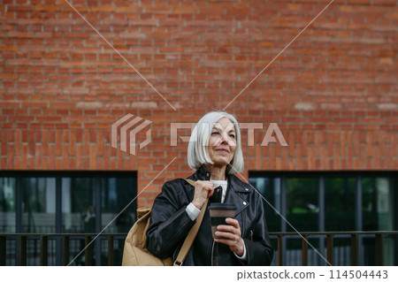 Portrait of stylish mature woman with gray hair on city street. Older woman in leather jacket with soft smile. 114504443