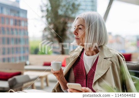 Attractive older woman checking smartphone, drinking coffee at coffee shop. Mature woman spending free time outdoors, in cafe, waiting for girl friends. 114504482