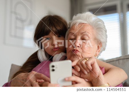 Grandmother with cute girl taking selfie with smartphone, making funny face. Portrait of an elderly woman spending time with granddaughter. 114504551