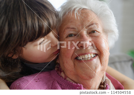 Cute girl hugging and kissing gradmother on cheek from behind. Portrait of an elderly woman spending time with granddaughter. 114504558
