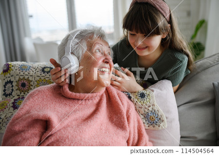Cute girl putting headphones on gradmother head. Portrait of an elderly woman spending time with granddaughter. Cute girl putting headphones on gradmother head. Portrait of an elderly woman spending time with granddaughter. 114504564