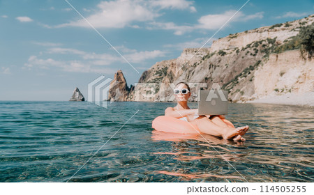 Woman freelancer works on laptop swimming in sea on pink inflatable ring. Happy tourist in sunglasses floating on inflatable donut and working on laptop computer in calm ocean. Remote working anywhere Woman freelancer works on laptop swimming in sea on pink inflatable ring. Happy tourist in sunglasses floating on inflatable donut and working on laptop computer in calm ocean. Remote working anywhere 114505255