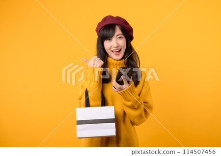 Happy young Asian woman in her 30s, holding a shopping paper bag and smartphone, celebrating her purchase with a cheerful smile, wearing a yellow sweater and red beret against a yellow background. Happy young Asian woman in her 30s, holding a shopping paper bag and smartphone, celebrating her purchase with a cheerful smile, wearing a yellow sweater and red beret against a yellow background. 114507456