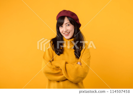 Happy young Asian woman in her 30s, wearing a yellow sweater and red beret, arms crossed on her chest, against a yellow background. Happy young Asian woman in her 30s, wearing a yellow sweater and red beret, arms crossed on her chest, against a yellow background. 114507458