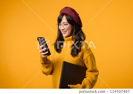 A young Asian woman, in a yellow sweater and red beret, confidently works on her laptop and smartphone against a vibrant yellow background. 114507467