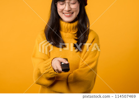 Close-up of a young Asian woman in her 30s, wearing a yellow sweater, happily changing channels on the TV with a remote control. Close-up of a young Asian woman in her 30s, wearing a yellow sweater, happily changing channels on the TV with a remote control. 114507469