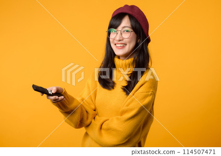 a comfortable home scene yellow background with a happy young woman in a yellow sweater, holding a remote control and watching TV. a comfortable home scene yellow background with a happy young woman in a yellow sweater, holding a remote control and watching TV. 114507471