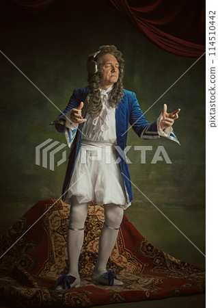 Elderly man dressed in richly detailed baroque costume with joyful expression listening music in headphones against vintage studio background. Elderly man dressed in richly detailed baroque costume with joyful expression listening music in headphones against vintage studio background. 114510442