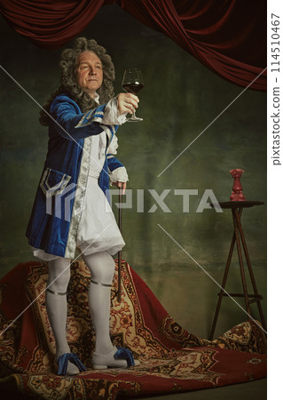 Elderly man wearing Baroque-style attire toasting and raising hand with glass of red wine against vintage studio background. Elderly man wearing Baroque-style attire toasting and raising hand with glass of red wine against vintage studio background. 114510467