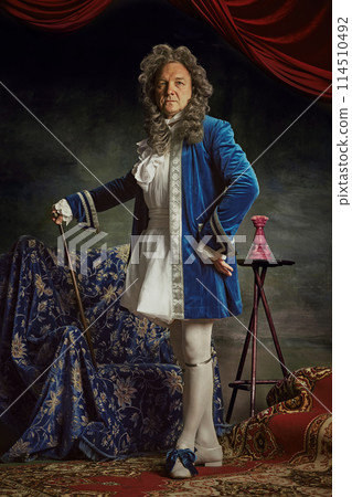 Full-length portrait of elderly man dressed as medieval person, aristocrat with confident expression against vintage studio background. Full-length portrait of elderly man dressed as medieval person, aristocrat with confident expression against vintage studio background. 114510492