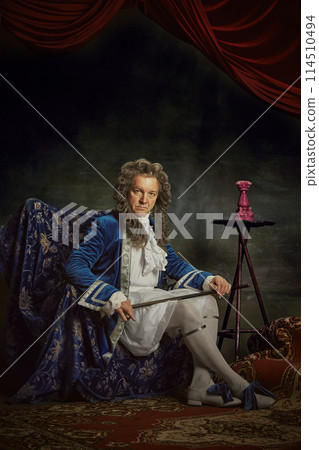 Portrait of elderly man, looks as medieval person, aristocrat, sitting on regally chair against vintage studio background. Portrait of elderly man, looks as medieval person, aristocrat, sitting on regally chair against vintage studio background. 114510494
