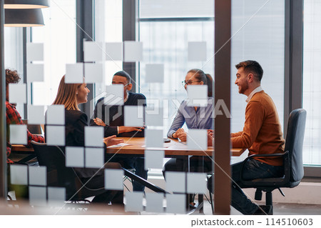 In a sleek glass office space, a team of business professionals engage in lively discussions and exchange strategic ideas, showcasing collaboration and innovation at the heart of modern corporate 114510603