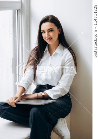 Attractive female freelancer sitting on windowsill with laptop. Woman preparing for working Attractive female freelancer sitting on windowsill with laptop. Woman preparing for working 114511228