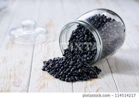 Raw black beans spill out of a glass storage jar on a white wooden table. Raw black beans spill out of a glass storage jar on a white wooden table. 114514217