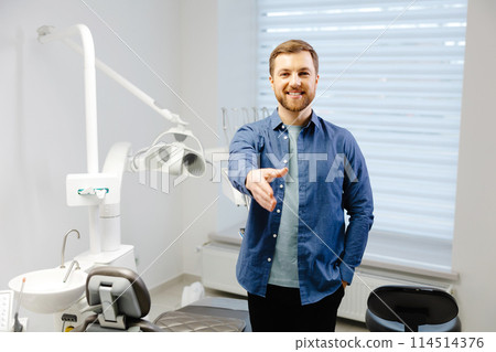 The happy adult man extends a hand of greetings, standing near stomatology chair in dentist office The happy adult man extends a hand of greetings, standing near stomatology chair in dentist office 114514376