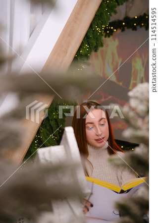 Hello winter. smiling modern woman in white sweater siting on chair and reading a book in room decorated for celebrating the new year and christmas festive mood 114514595
