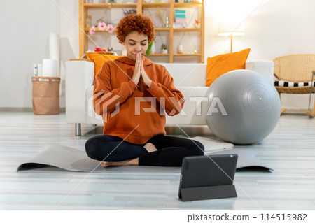 Yoga mindfulness meditation. Young healthy african girl practicing yoga at home. Woman sitting in lotus pose on yoga mat eyes close hands in prayer meditating indoor. Girl doing breathing practice 114515982