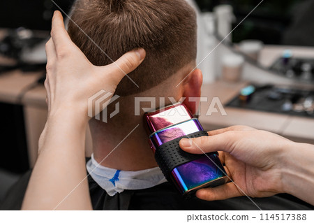 Barber uses shaver machine to cut young man hair in barbershop closeup. Master does modern haircut to male client in professional salon 114517388