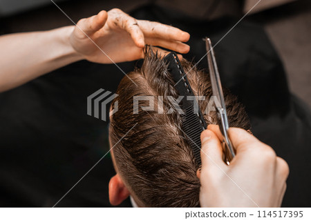Stylist cuts male client hair using comb and scissors in barbershop closeup. Barber with professional tools serves man in salon 114517395