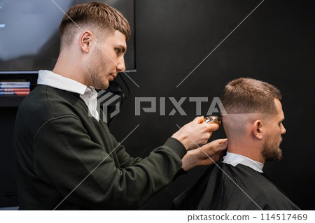 Young barber cuts mans hair using trimmer in the barbershop. Young barber cuts mans hair using trimmer in the barbershop. 114517469
