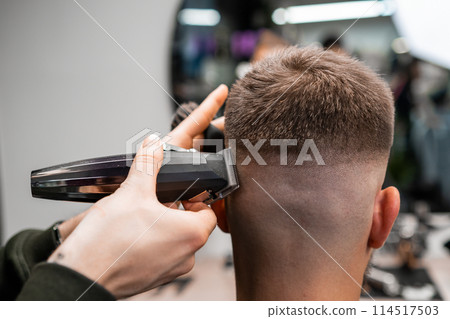 Skilled barber cuts client short hair with electrical trimmer in barbershop closeup. Hairdresser does stylish men haircut in beauty shop 114517503