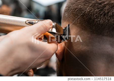 Coiffeur shaves client hair with electrical trimmer in modern barbershop closeup. Barber does elegant haircut contour in beauty salon Coiffeur shaves client hair with electrical trimmer in modern barbershop closeup. Barber does elegant haircut contour in beauty salon 114517518