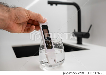 Man hand immerses conductometer in a beaker of water to check purity in front of a modern kitchen water faucet. Conductometer shows that water is dirty  114517633