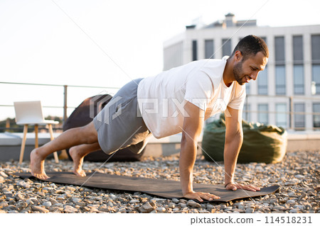 Young smiling sports man doing morning exercises on roof of modern house. 114518231