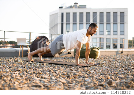 Young smiling sports man doing morning exercises on roof of modern house. 114518232
