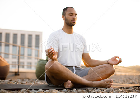 Male meditating in lotus position while sitting on mat on roof of modern house. 114518237