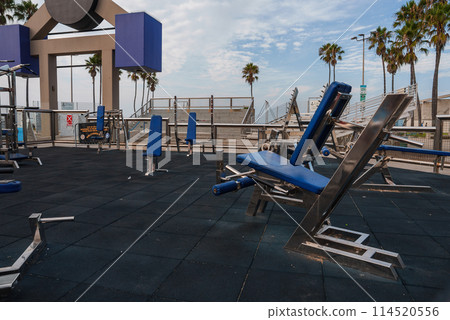 Outdoor gym area with various fitness equipment like weight benches and pull up bars. Located at Venice Beach, with palm trees and a cloudy sky in the background. Outdoor gym area with various fitness equipment like weight benches and pull up bars. Located at Venice Beach, with palm trees and a cloudy sky in the background. 114520556