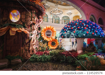 Vibrant indoor scene in Las Vegas Strip hotel with whimsical tree, colorful floral mushroom, and lush garden. Luxurious decor and lively visitors add to enchanting ambiance. 114520574