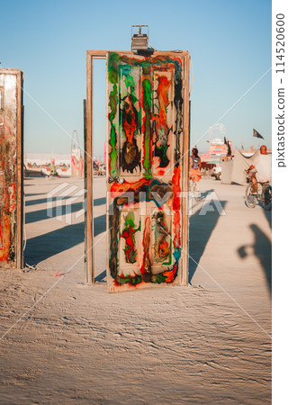 Vibrant art installation with abstract colors in desert festival setting. Adorned structure of metallic frame against blue sky, attendees in distance. 114520600