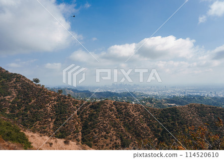 Scenic view of arid hills surrounding Los Angeles with a cityscape in the distance under a partly cloudy sky. Helicopter adds dynamism to the tranquil scene. 114520610