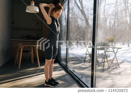 Full length portrait of athletic young woman in outfit preforming warming up exercise, stretching muscles before workout indoors, standing by window with snowy winter nature view on sunny day. 114520872