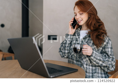 Smiling female manager holding talking on smartphone, working on laptop, drinking coffee sitting at desk in cozy living room. Multitasking concept, home office, remote work or e-learning. 114520922