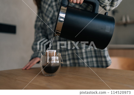 Close-up cropped shot of pouring hot water to make coffee with drip coffee bag on table. Hot water poured into cup of roasted drink to produce fresh drip coffee. Making coffee with drip bag 114520973