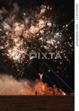 Spectacular desert event at night with fireworks, a wooden structure with illuminated human figure, and silhouettes of onlookers in the foreground. Celebratory atmosphere. 114520989