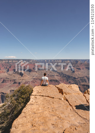 Person sitting on edge of rock formation, gazing over grand canyon with layers of red, brown, and orange sedimentary rocks. Clear blue sky, hinting at Grand Canyon, USA. Person sitting on edge of rock formation, gazing over grand canyon with layers of red, brown, and orange sedimentary rocks. Clear blue sky, hinting at Grand Canyon, USA. 114521030